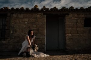 Portrait de mariée dramatique - Ruines des Bardenas au coucher du soleil Mariée en robe blanche assise avec bouquet et chapeau devant une maison en ruine aux Bardenas, éclairée par la lumière dorée du soir